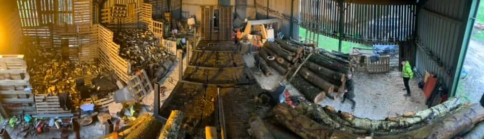 Wide view of the T & H Logs barn with sawmill, log piles and workers in hi-vis