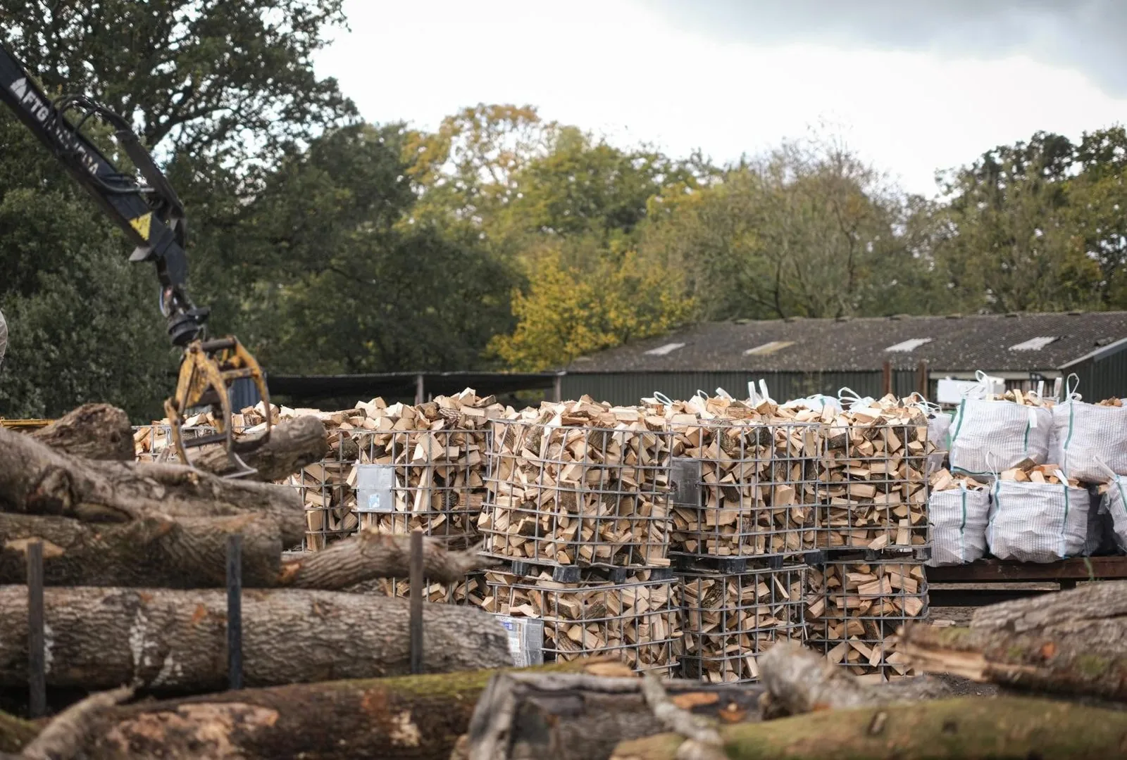 A crane grab over the timber pile, beside crates of split logs and bags.