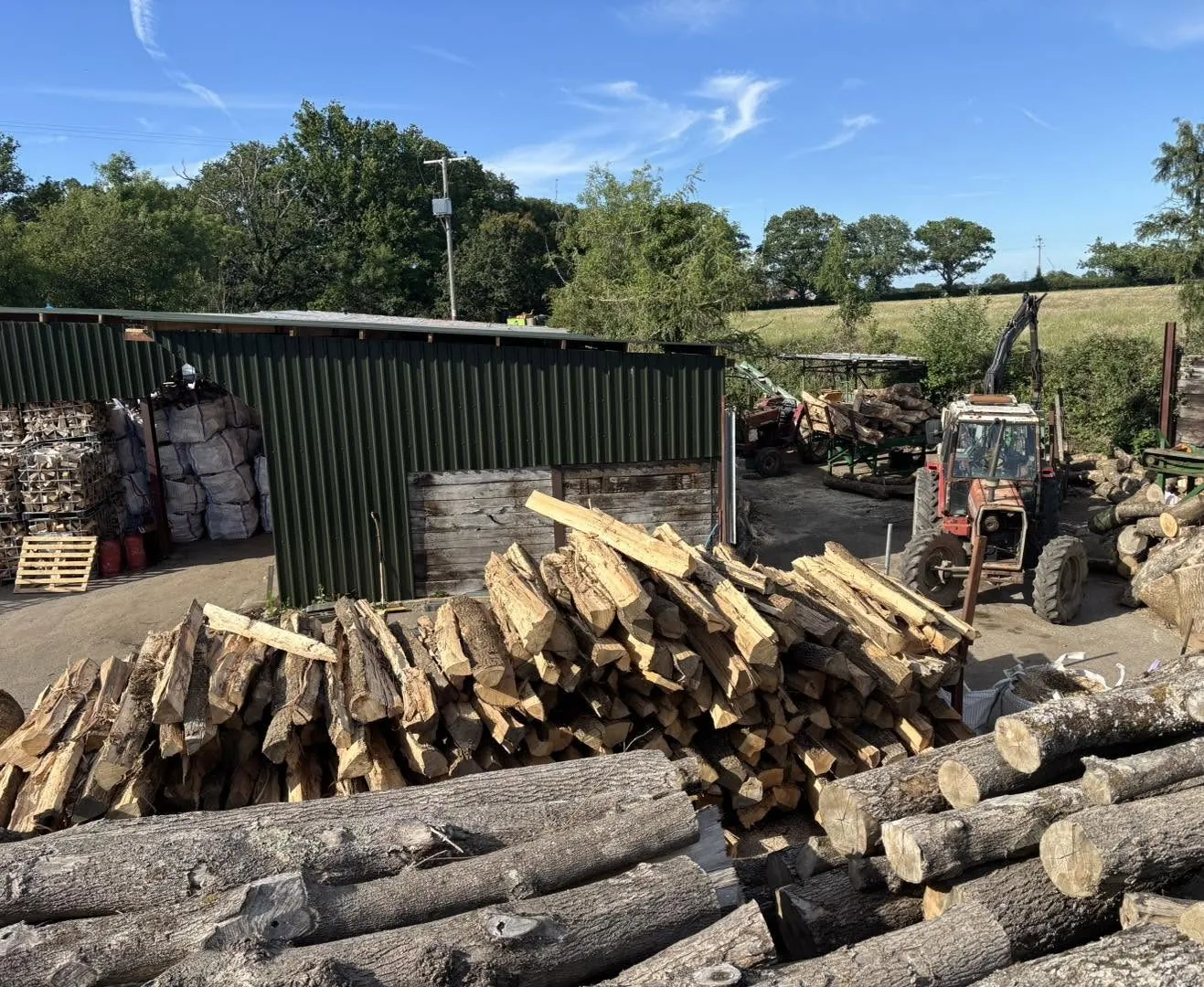 A pile of cut logs beside a tractor, with bagged firewood behind.
