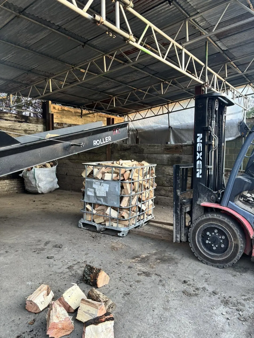 A forklift beside a roller conveyor and a crate of split logs under cover.