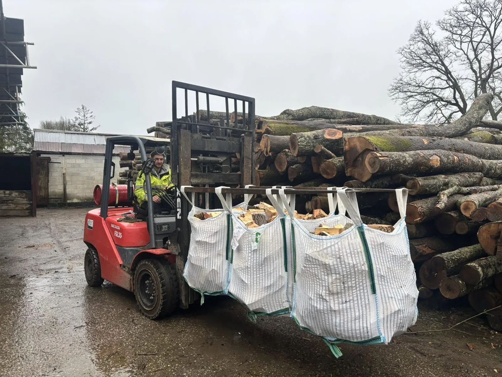 A forklift carries bags of split logs past a large pile of round logs.
