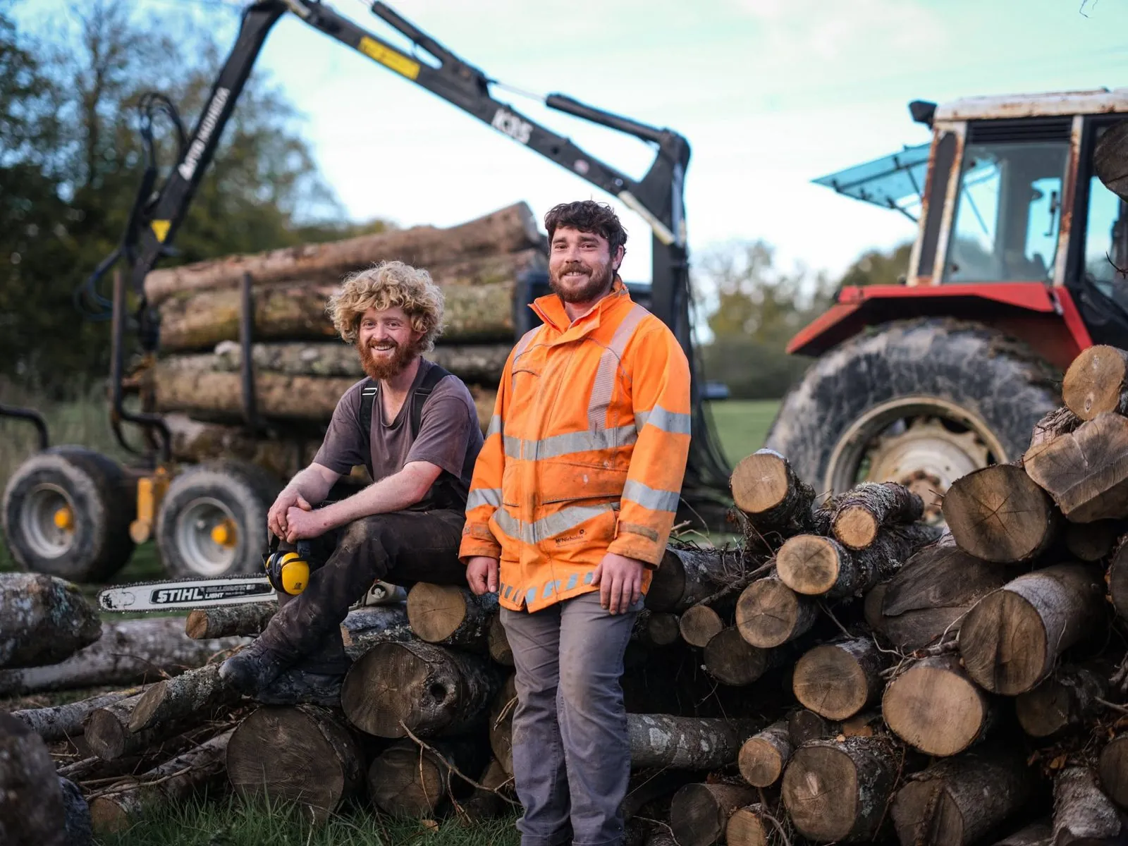 Two of the team resting on a log pile, with a tractor and crane trailer behind.