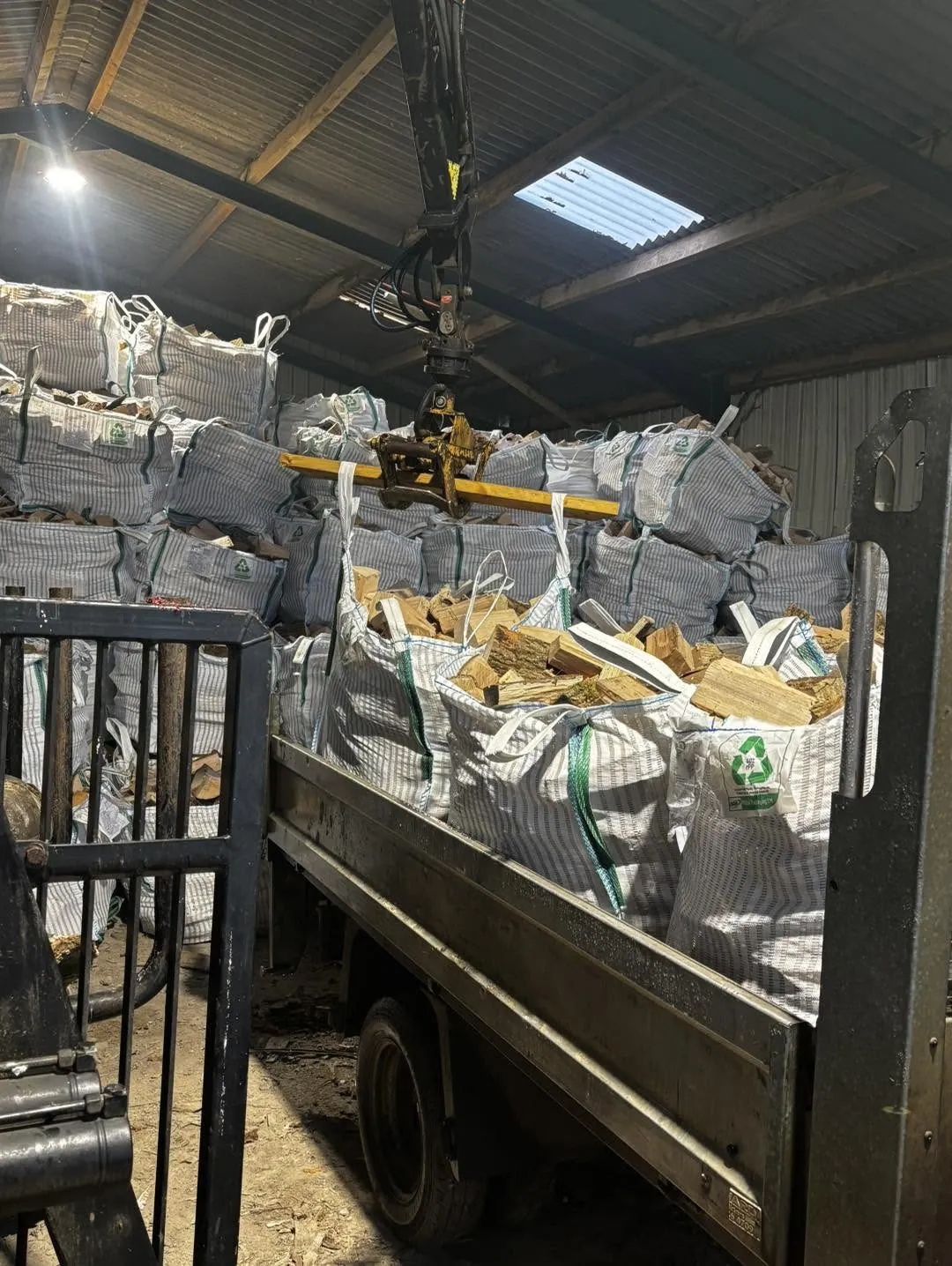 A truck loaded with bags of logs inside a barn, with a crane grab overhead.
