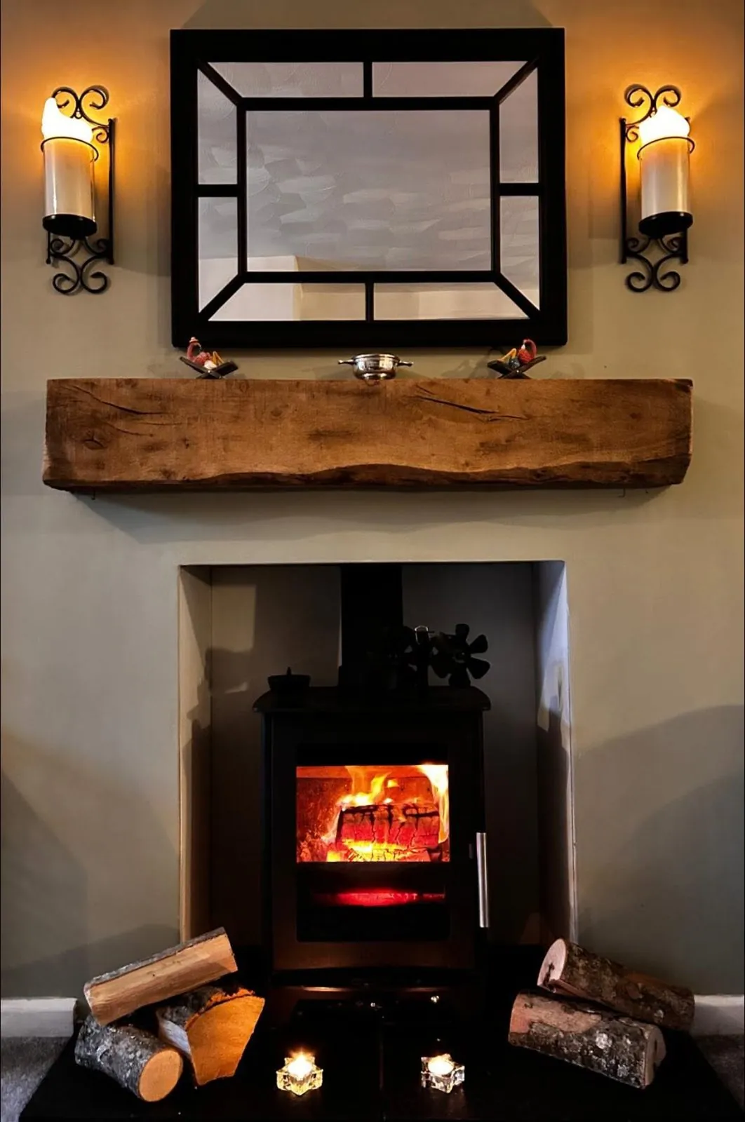 A wood burner alight in a fireplace, with logs beside it and a mirror above.